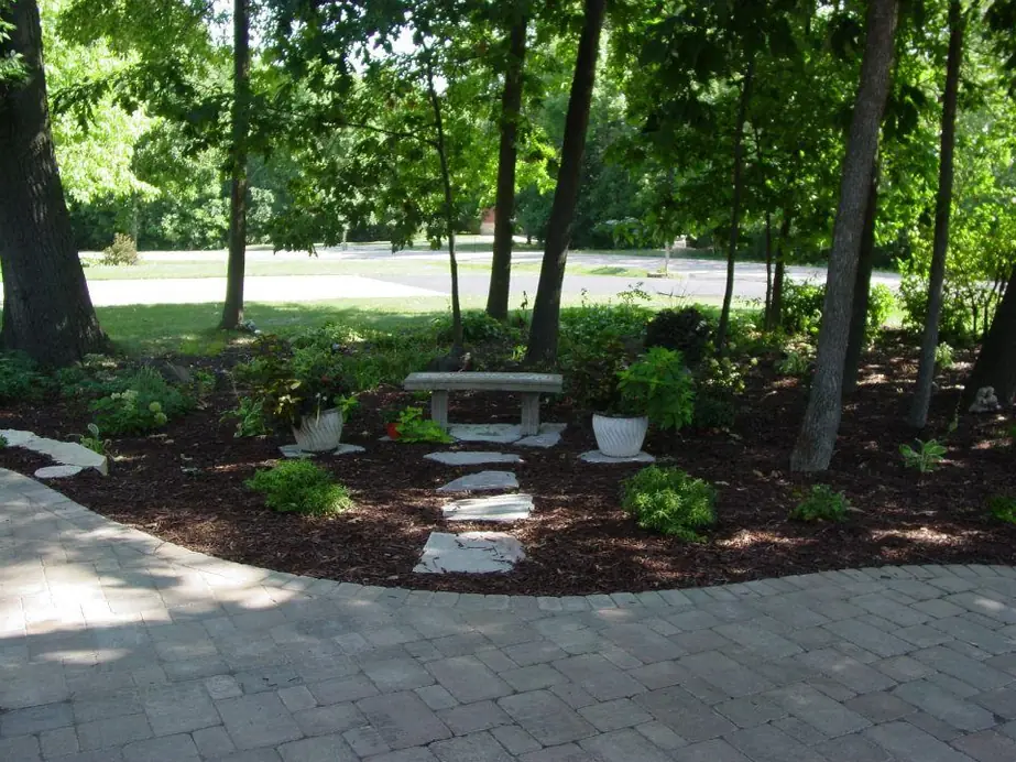 A stone bench on flagstone steppers set into hardwood bark beds under mature oaks, off a paver patio on an Oshkosh property